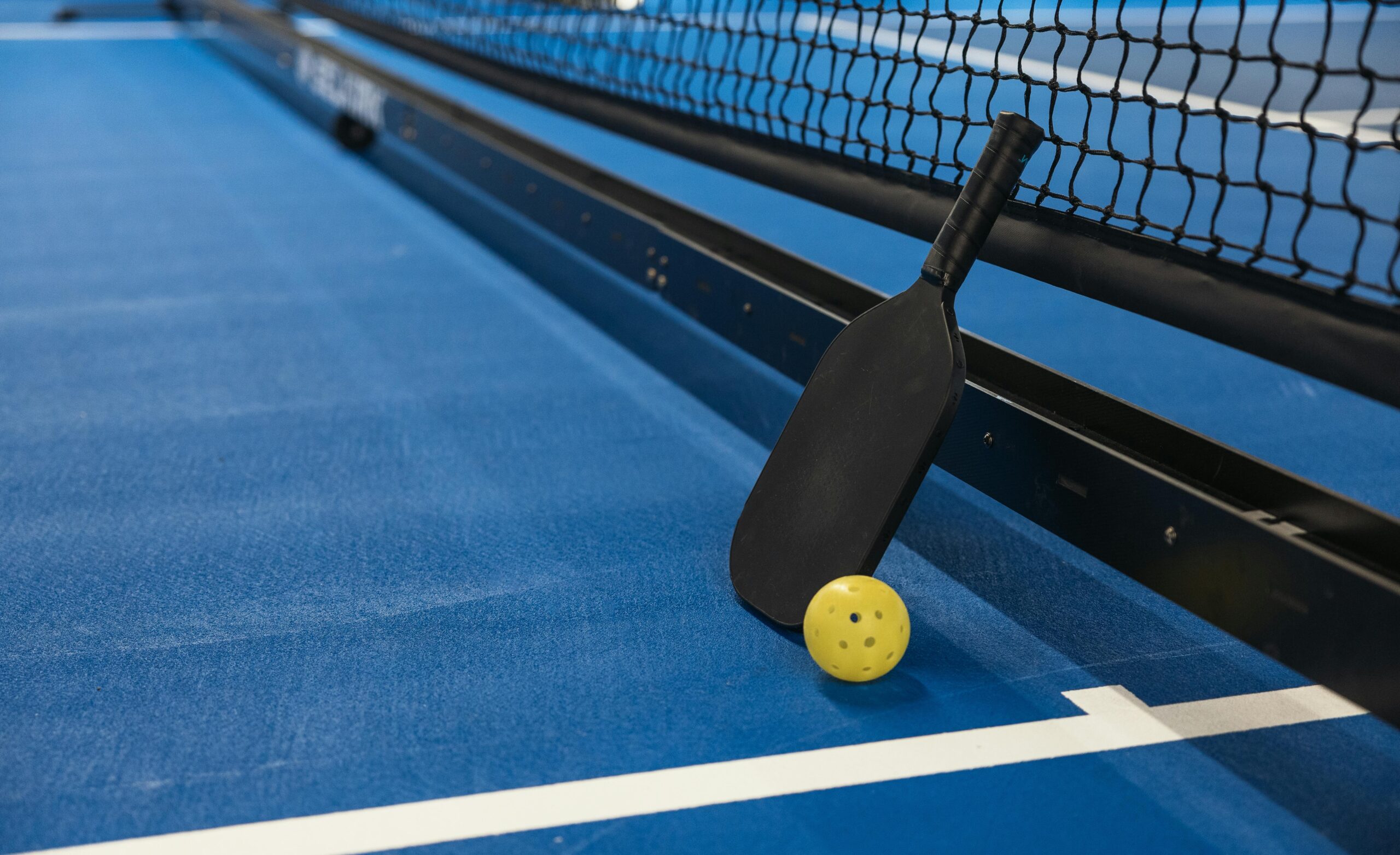 A black pickleball paddle and a yellow perforated ball resting against the net on a blue indoor pickleball court.