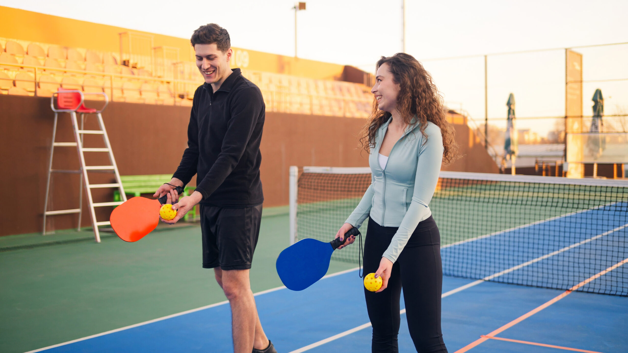 Young adults playing pickleball on an outdoor court, holding paddles and a perforated ball, smiling and enjoying a sunny day